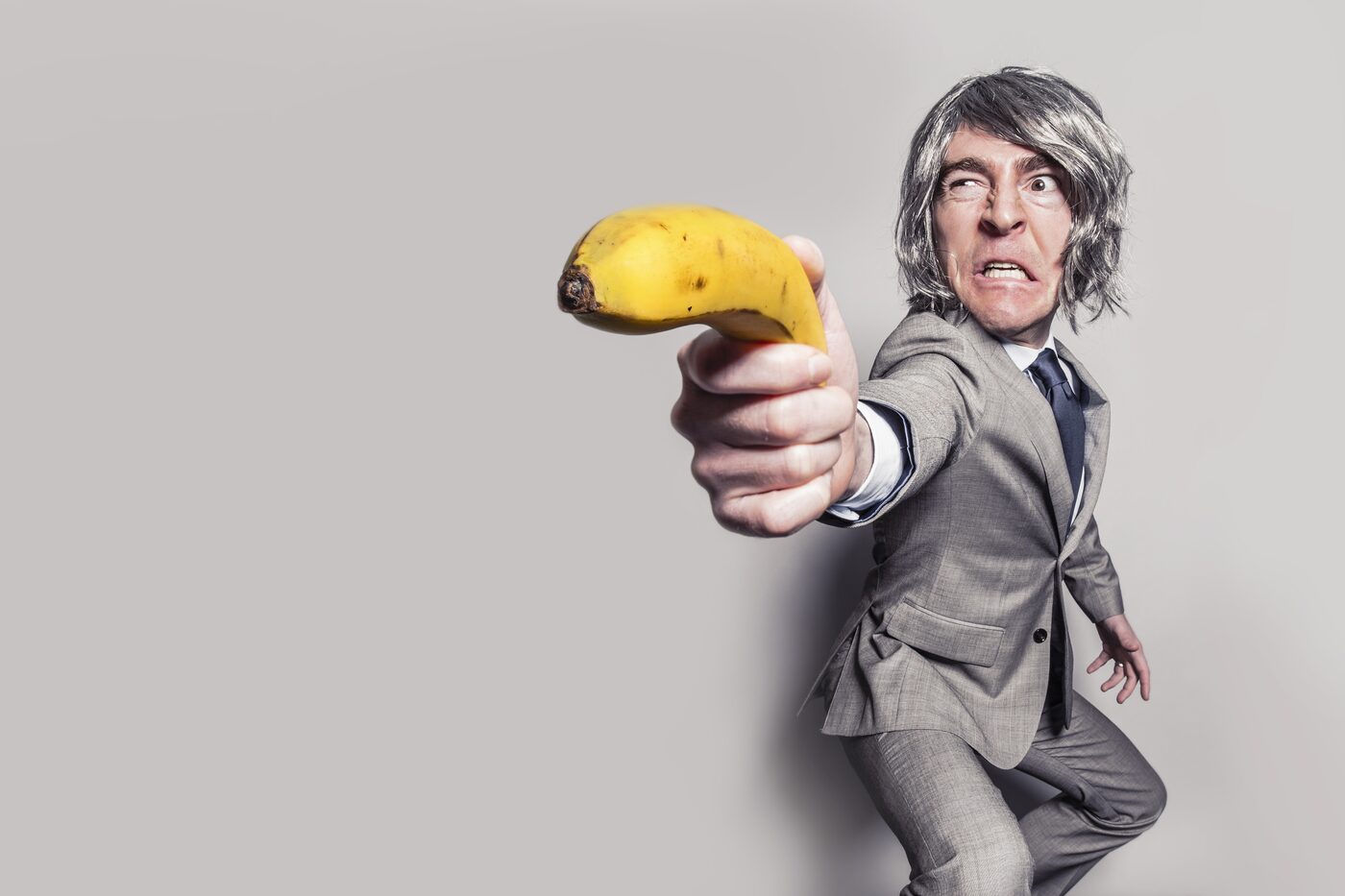 Mann im Anzug mit grauer Perücke zielt grimmig mit einer Banane wie mit einer Pistole, humorvolle Inszenierung