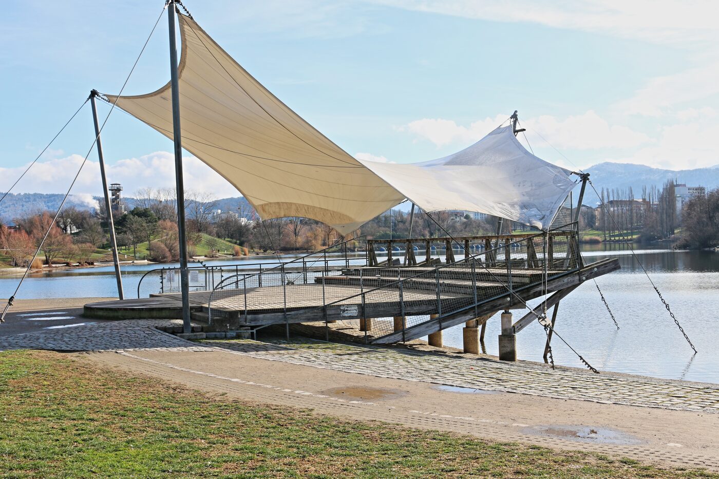 Überdachte Seebühne mit segelartigen Sonnenschutz-Membranen an einem See, Berge im Hintergrund
